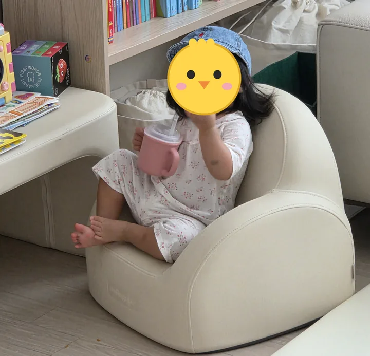크림색 아기 소파에 기대 앉아 분홍 컵을 든 첫째. 옆에 같은 색 아기 책상이 보인다(얼굴은 병아리 그림으로 가림)