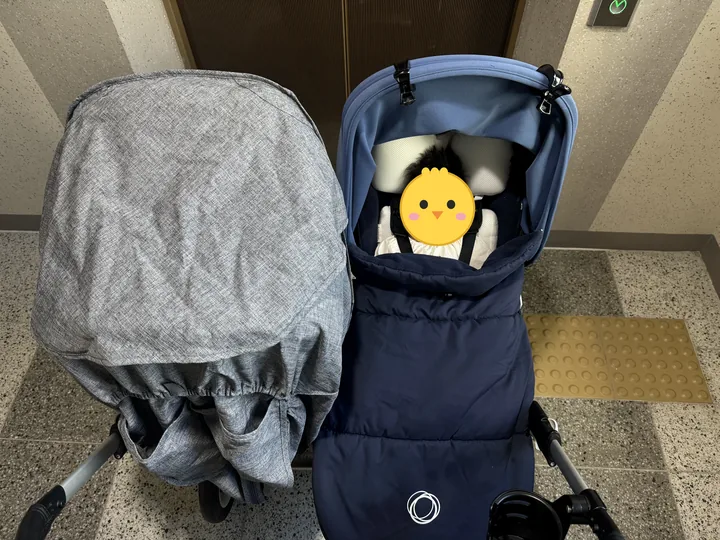 엘리베이터 앞 쌍둥이 유모차. 왼쪽 칸은 회색 방풍커버, 오른쪽 칸은 네이비 부가부 풋머프 속 둘째(얼굴은 병아리 그림으로 가림)