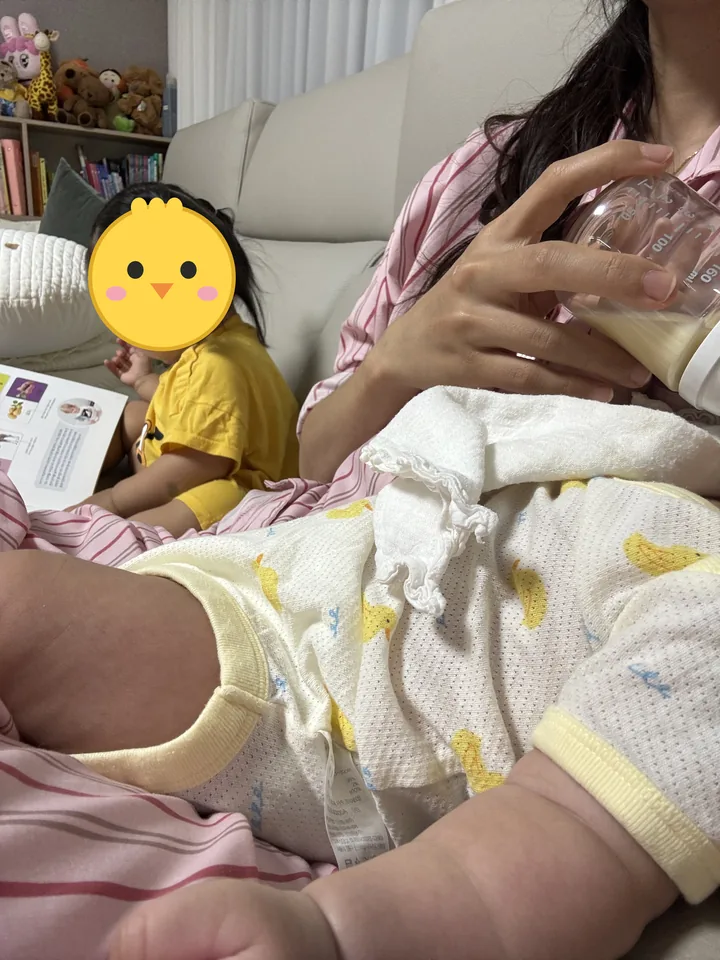 소파에서 둘째에게 분유를 먹이는 엄마와 옆에서 책을 보는 첫째(첫째 옆얼굴은 병아리 그림으로 가림)