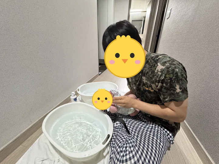 복도 바닥에 대야 두 개를 놓고 신생아 첫째를 목욕시키는 아빠(얼굴은 병아리 그림으로 가림)