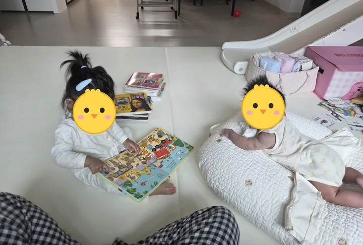 매트 위에서 그림책을 펼친 첫째와 수유쿠션에 엎드려 터미타임 중인 둘째(두 아이 얼굴은 병아리 그림으로 가림)