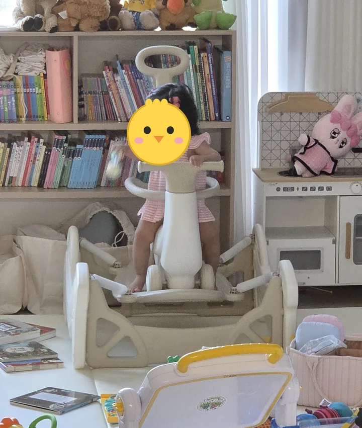 분홍 줄무늬 옷을 입은 첫째가 거실 책장 앞에서 흔들 받침 위 베이지 스프링카 손잡이를 잡고 서 있다(얼굴은 병아리 그림으로 가림)